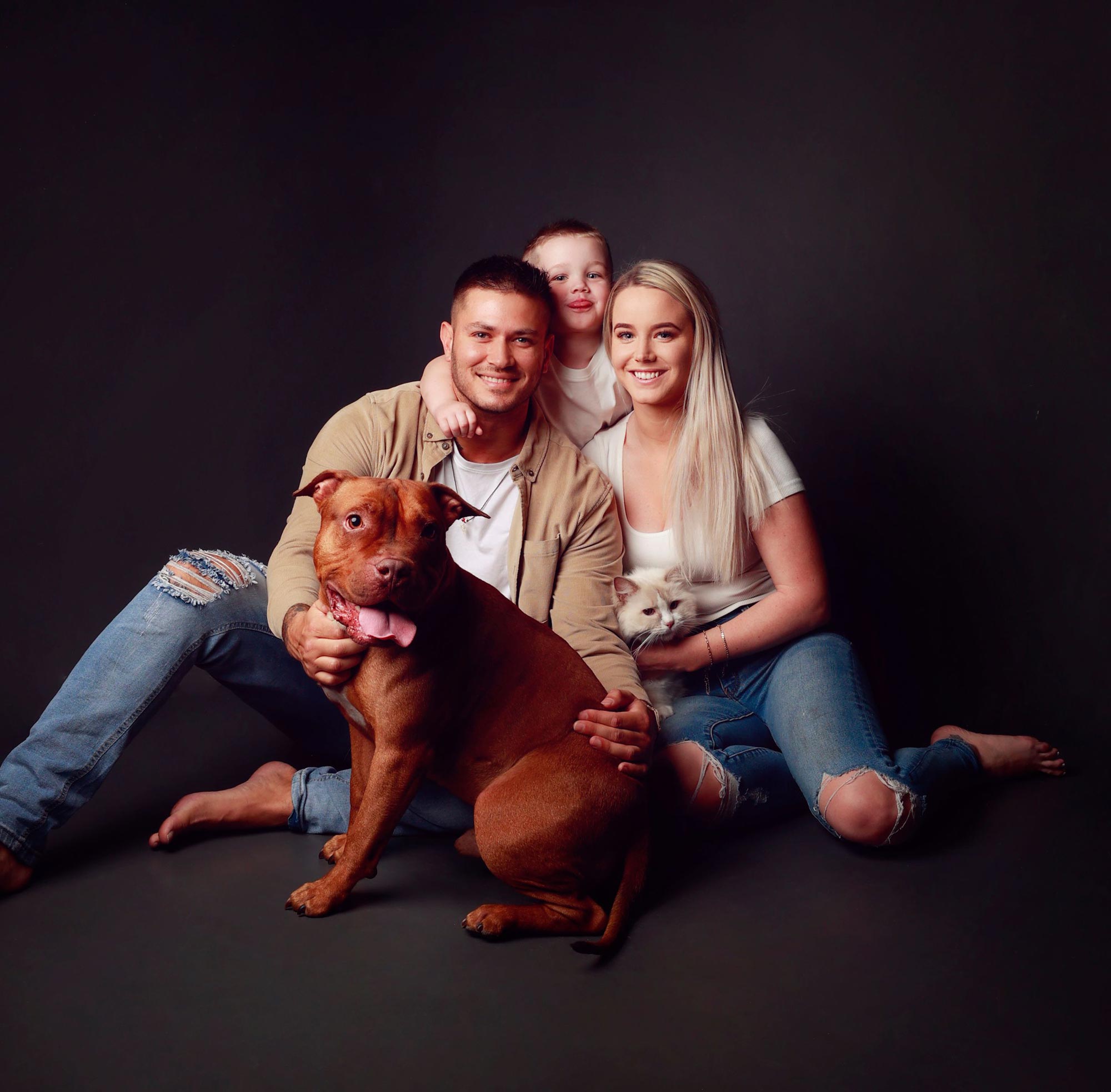 A young family sitting together on the studio floor with their pets, on a dark backdrop