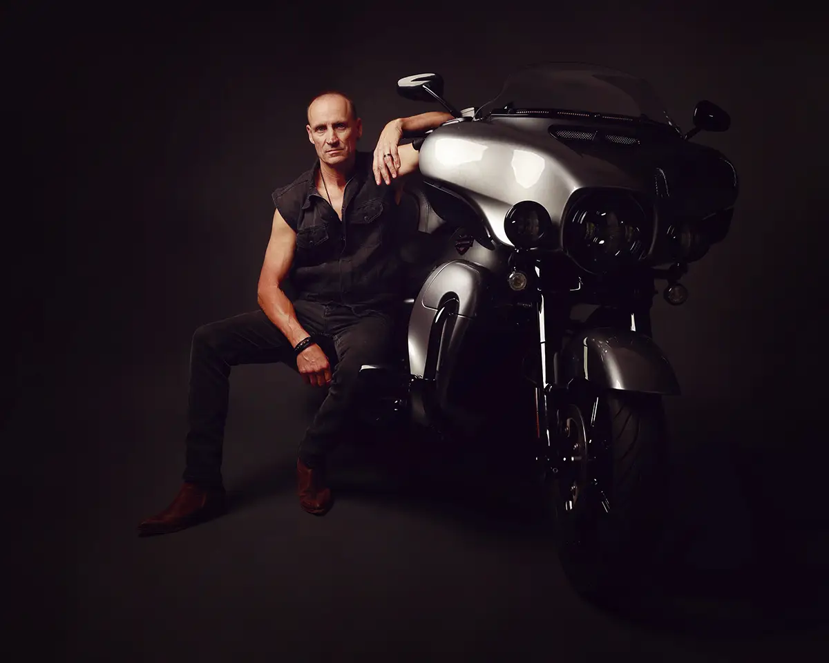A rider leaning against his motorbike, lit on a dark studio backdrop