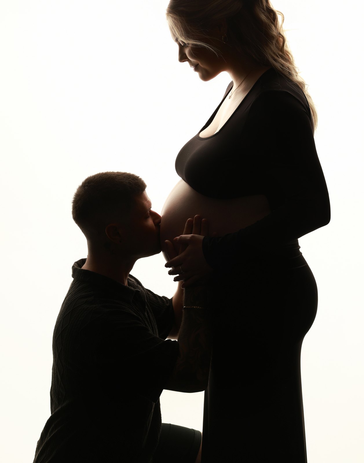 An expectant couple in silhouette, her partner kneeling to kiss the bump